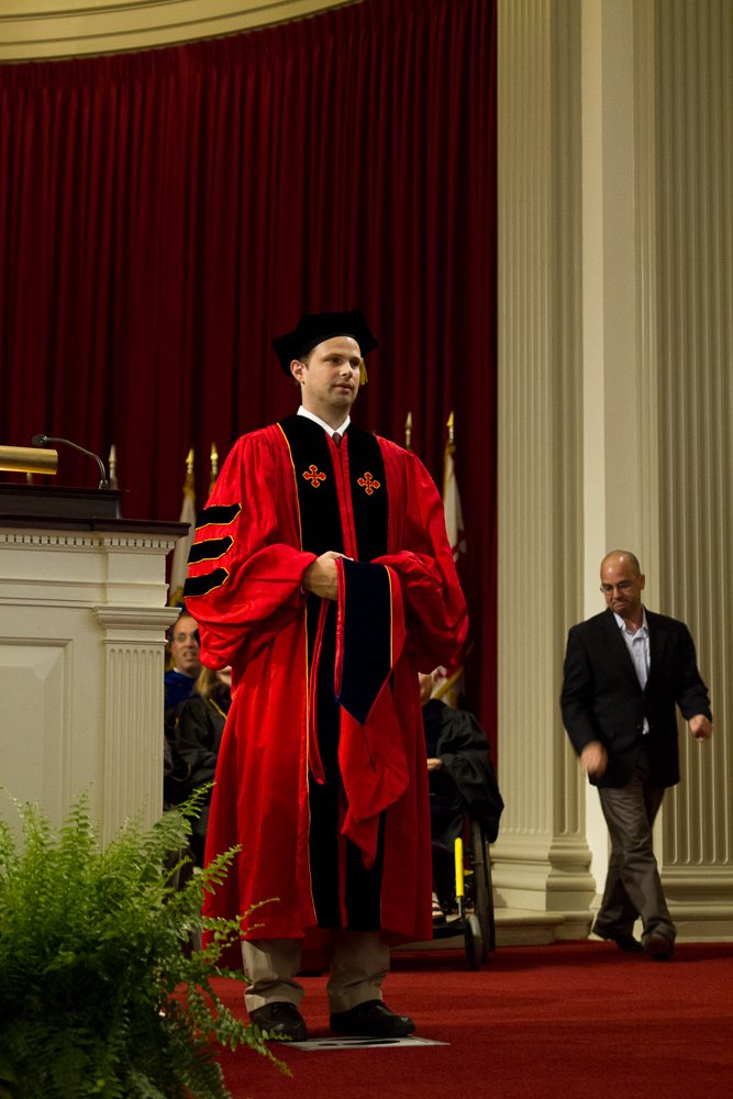Robert stands in Phd regalia as advisor walks behind.